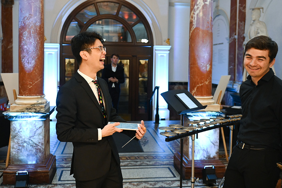 Two students laughing in the RCM's entrance hall, one holding an iPad. A percussion instrument and another student are behind them.